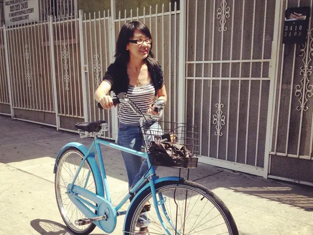 In this image in the center there is one woman who is holding a cycle and walking, and there is one...